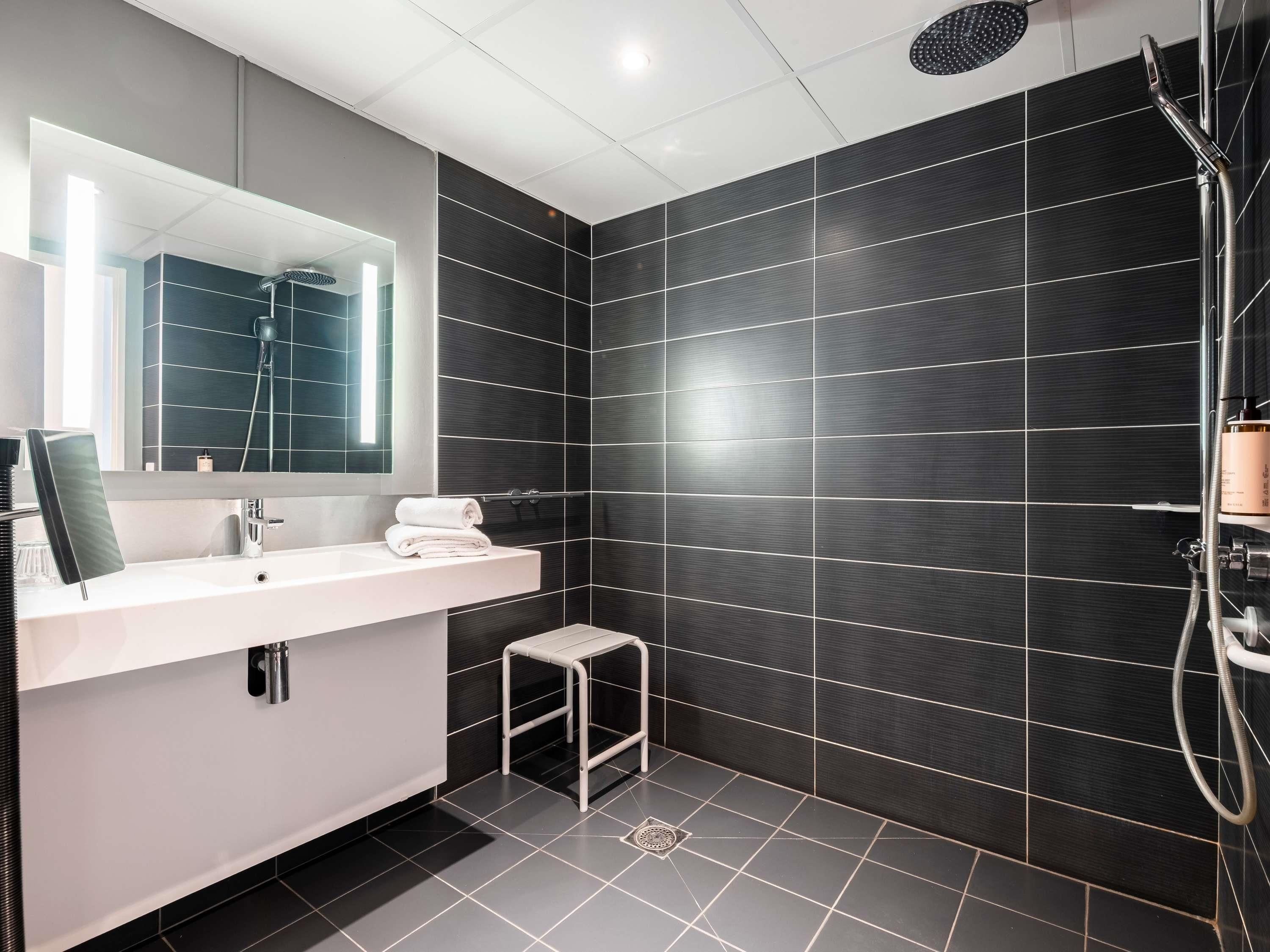 Une salle de bain moderne avec un grand miroir, un lavabo blanc et des murs recouverts de carreaux foncés.
