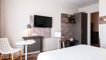 A hotel room with a bed, a desk with a lamp, a chair, a TV mounted on the wall, and a small table with a handbag.