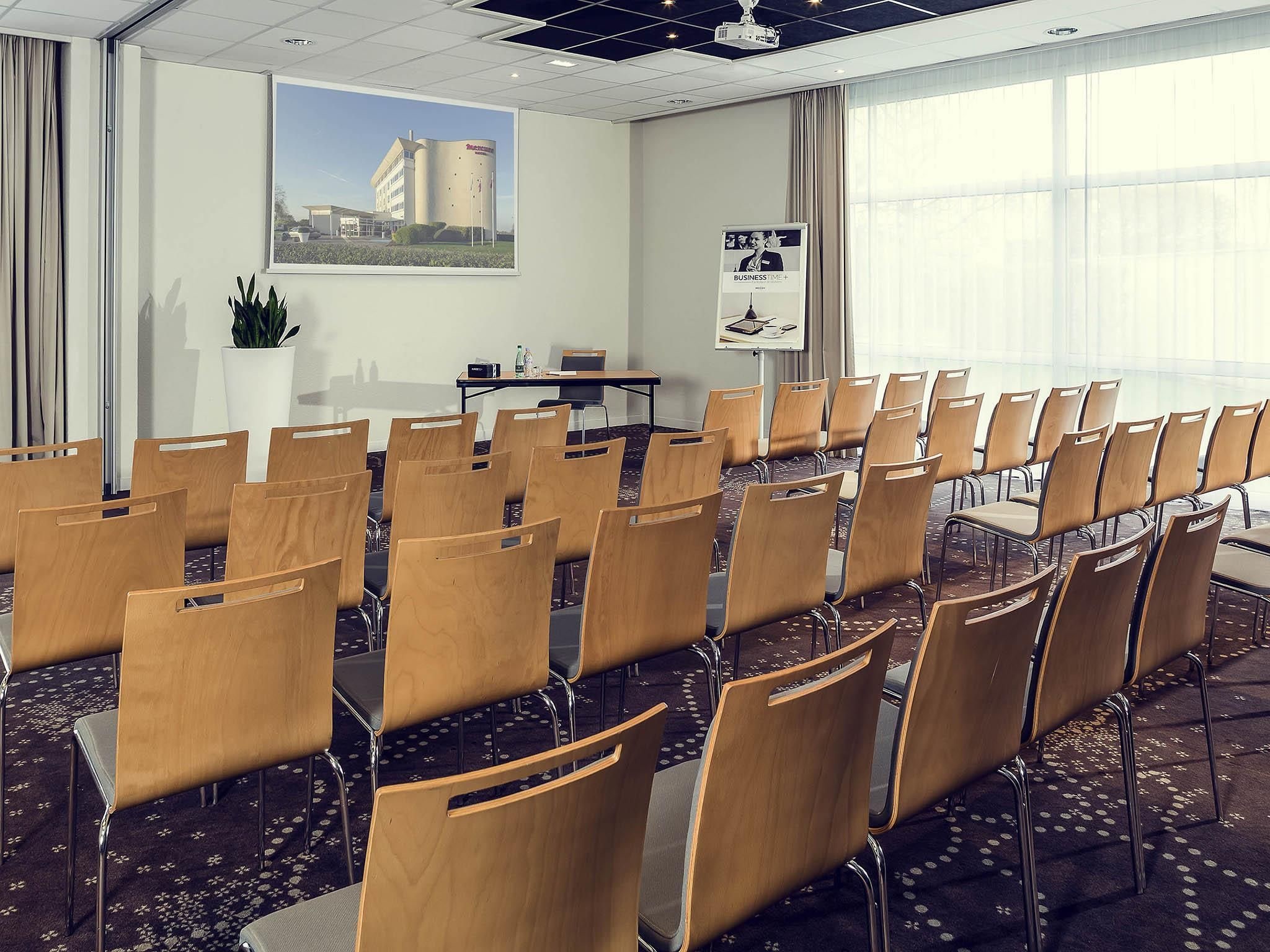 Un salón de conferencias con filas de sillas de madera, una pantalla de proyector, una mesa con una computadora portátil y un amplio ventanal con vista a un edificio.