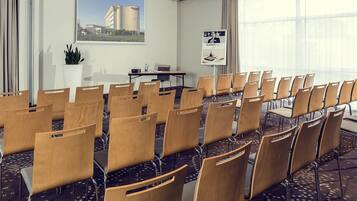 Un salón de conferencias con filas de sillas de madera, una pantalla de proyector, una mesa con una computadora portátil y un amplio ventanal con vista a un edificio.