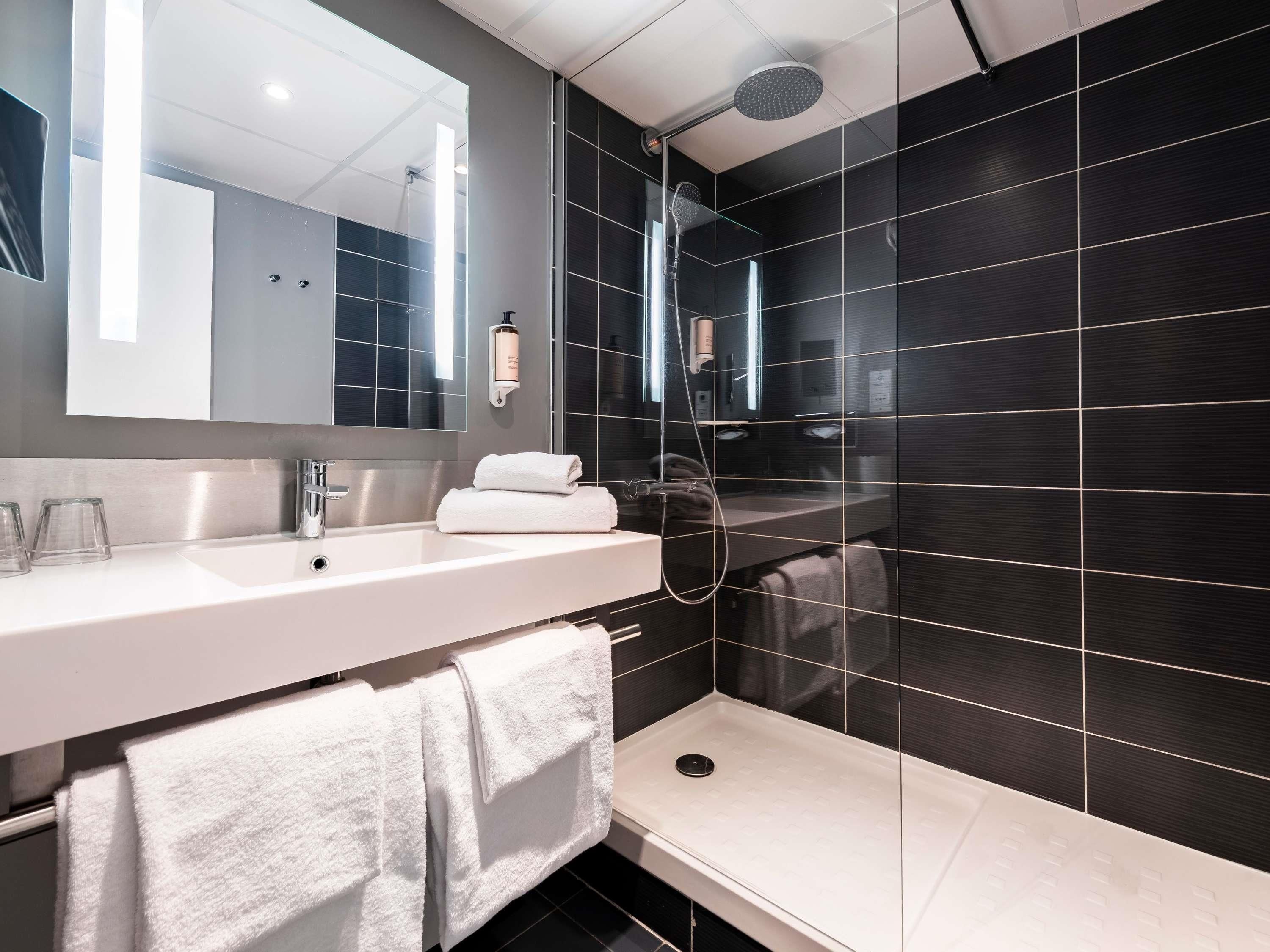 A modern bathroom with a large mirror, a white sink, a shower with a handheld showerhead, and dark tiled walls.