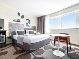 Quarto de hotel moderno com cama bem arrumada, uma pequena mesa redonda e vista do céu através de uma grande janela.