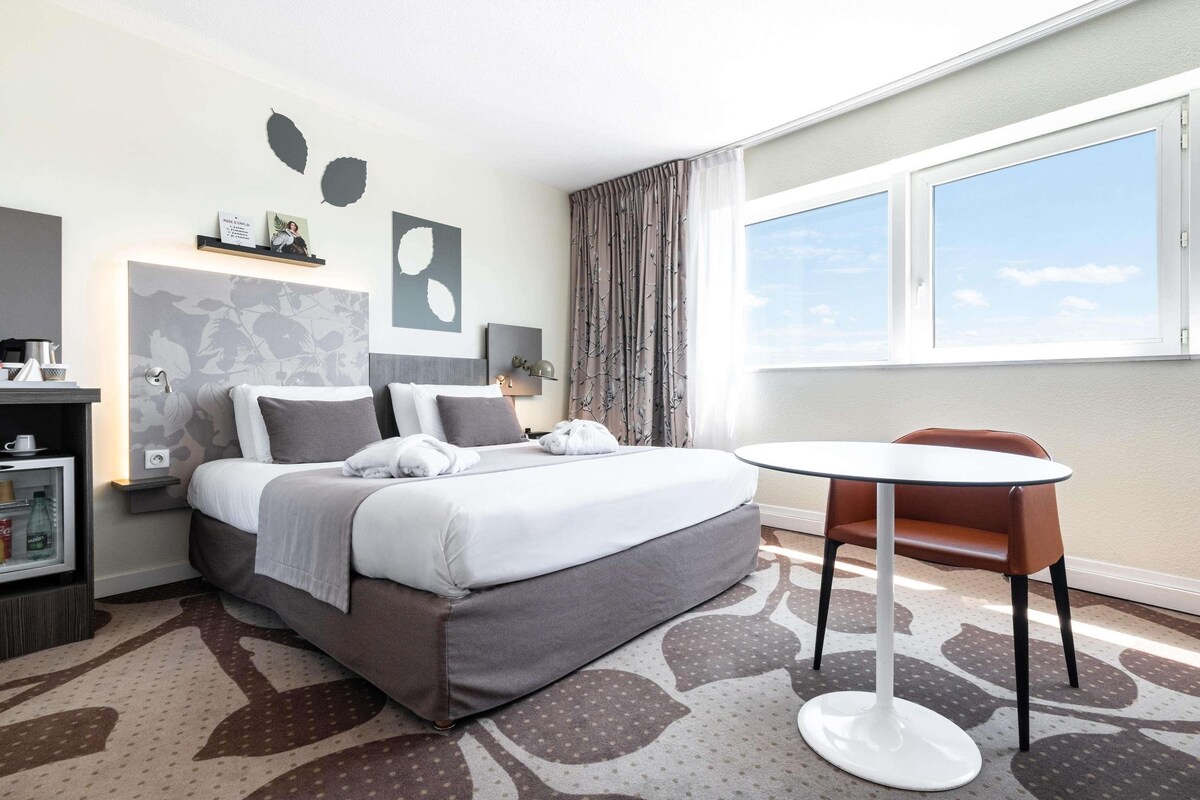 A hotel room with a bed, a bedside table, a chair, a window, and a view of the sky.