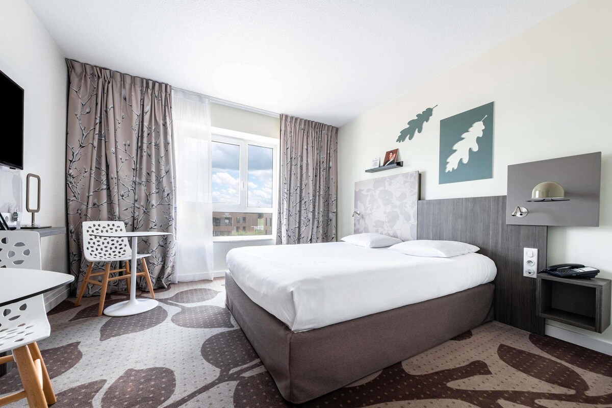 A hotel room with a bed, a desk, a chair, a TV, and a window with curtains.