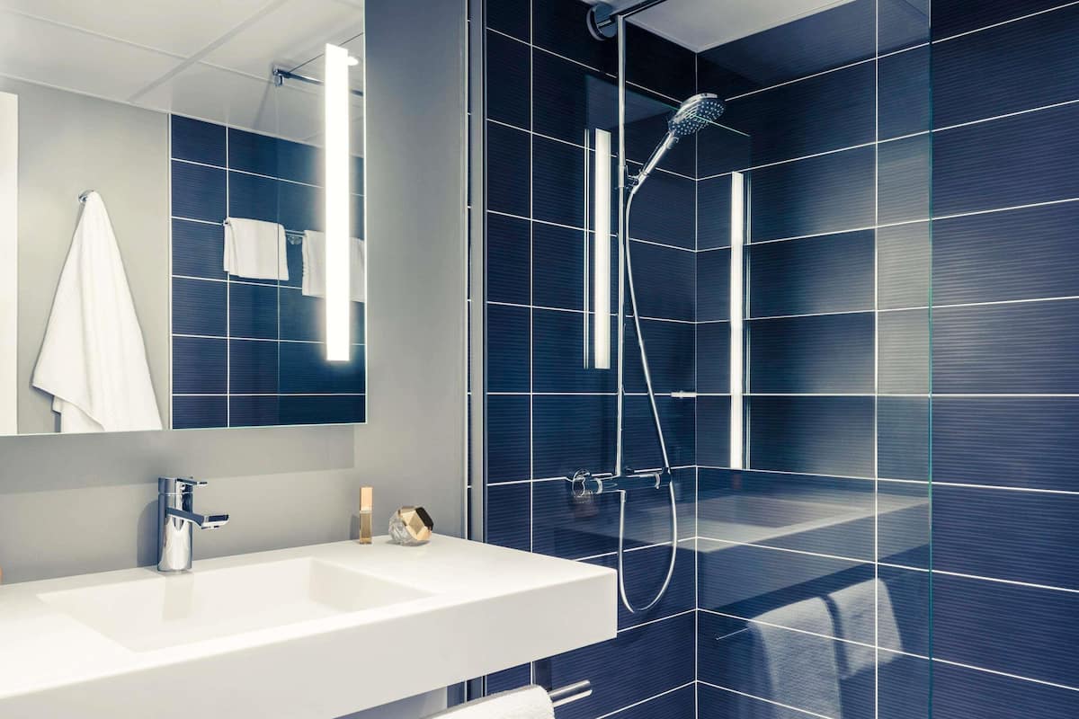 A modern bathroom with a large mirror, a white sink, a handheld showerhead, and dark blue tiled walls.