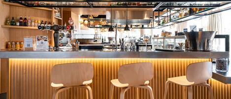 A bar area with wooden stools, shelves of bottles, and a kitchen in the background.
