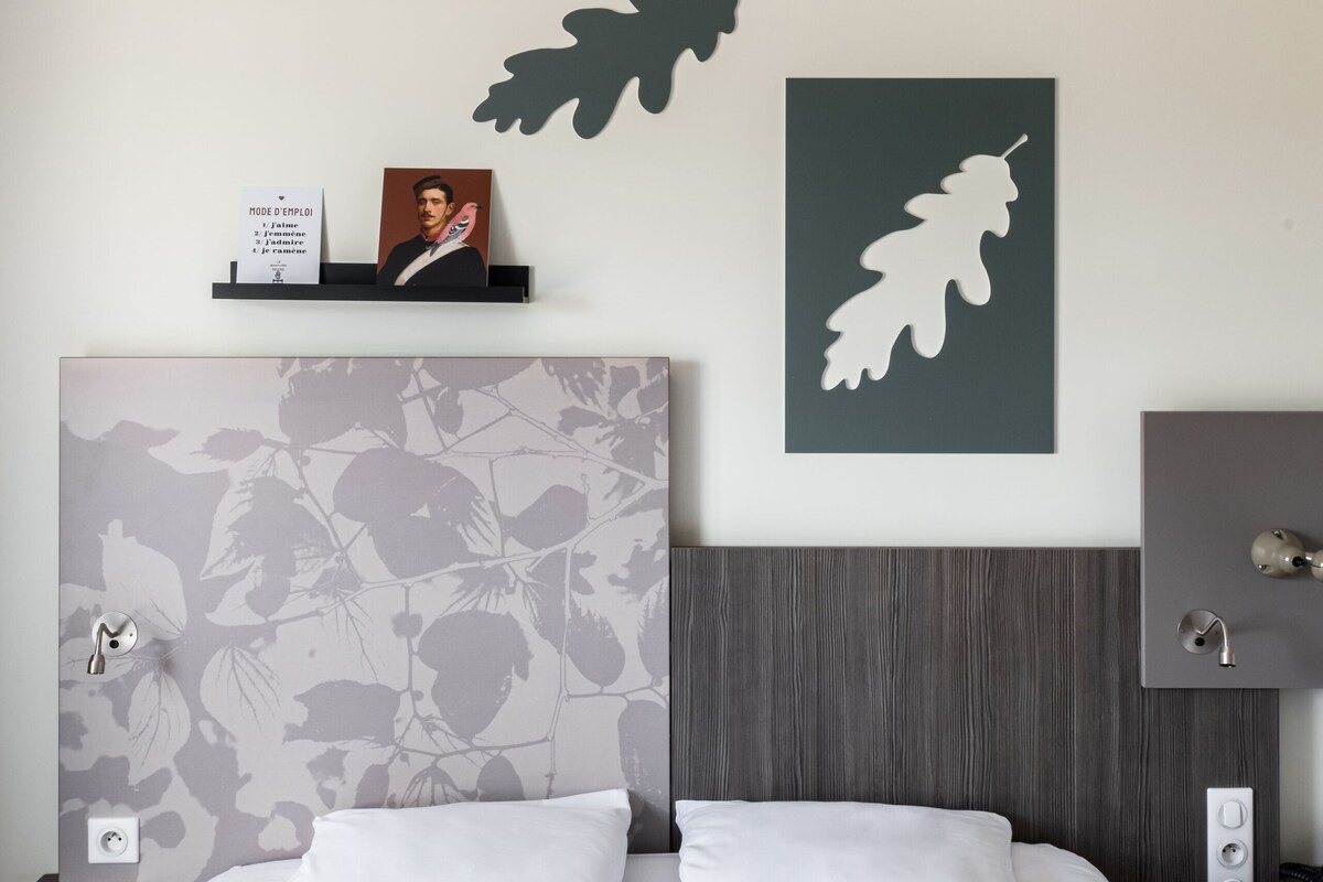 A hotel room with a large bed, a headboard with a floral pattern, a wall-mounted shelf with a framed photo and a sign, and a wall art piece of a leaf.