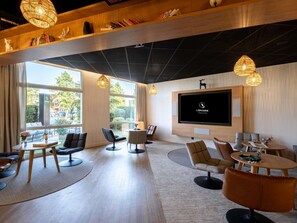 Un moderno vestíbulo de hotel con un televisor grande, asientos cómodos, una estantería y ventanas amplias.