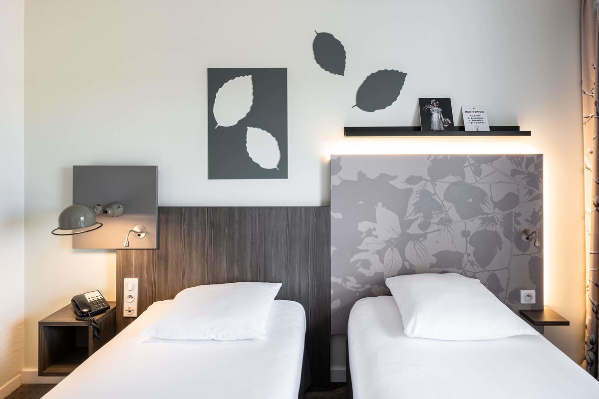 A hotel room with two beds, a desk with a phone, a wall-mounted lamp, and artwork.
