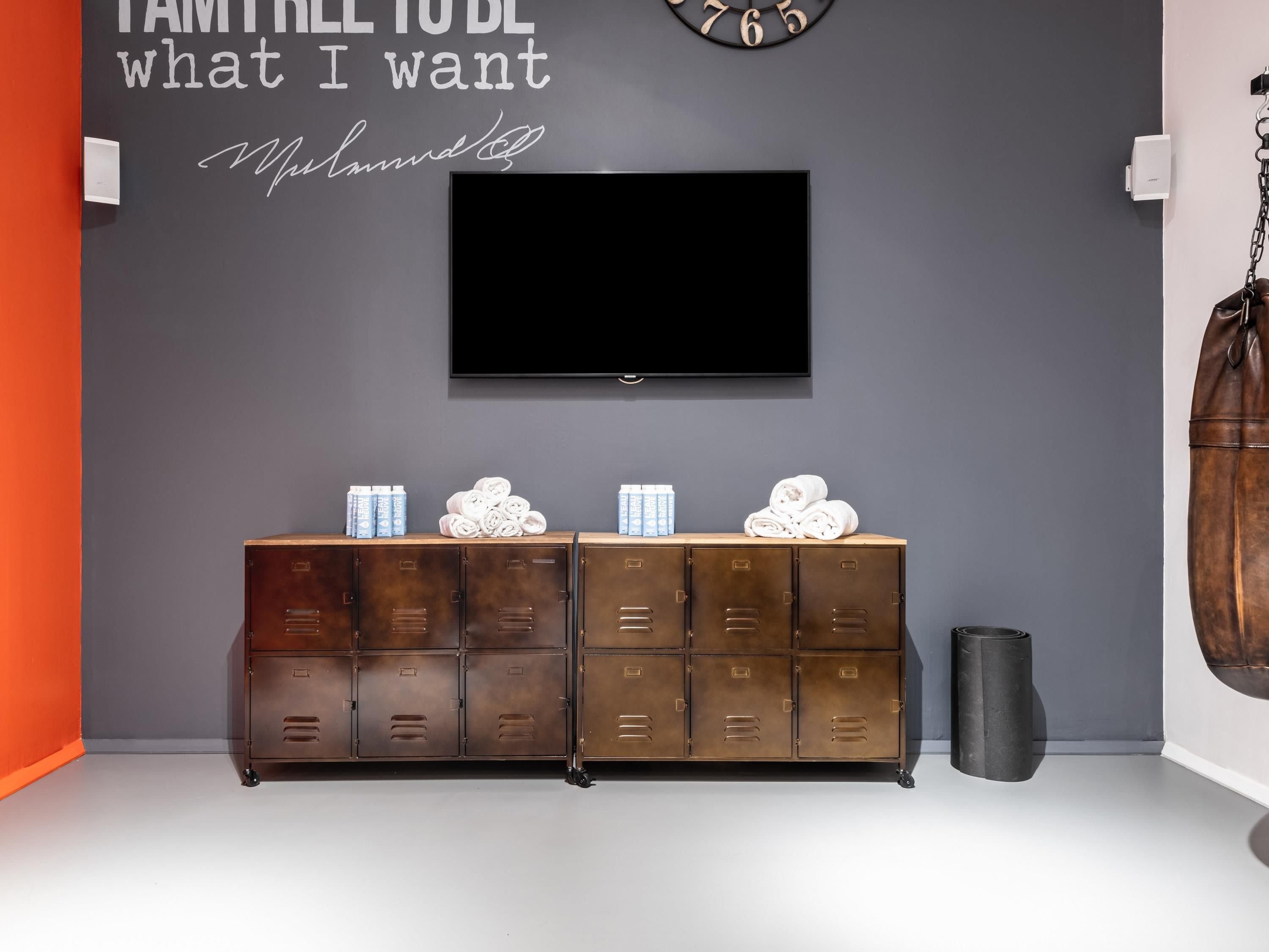 A room with a TV mounted on the wall, a wooden cabinet with towels and toiletries, and a clock.