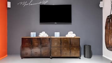 A room with a TV mounted on the wall, a wooden cabinet with towels and toiletries, and a clock.