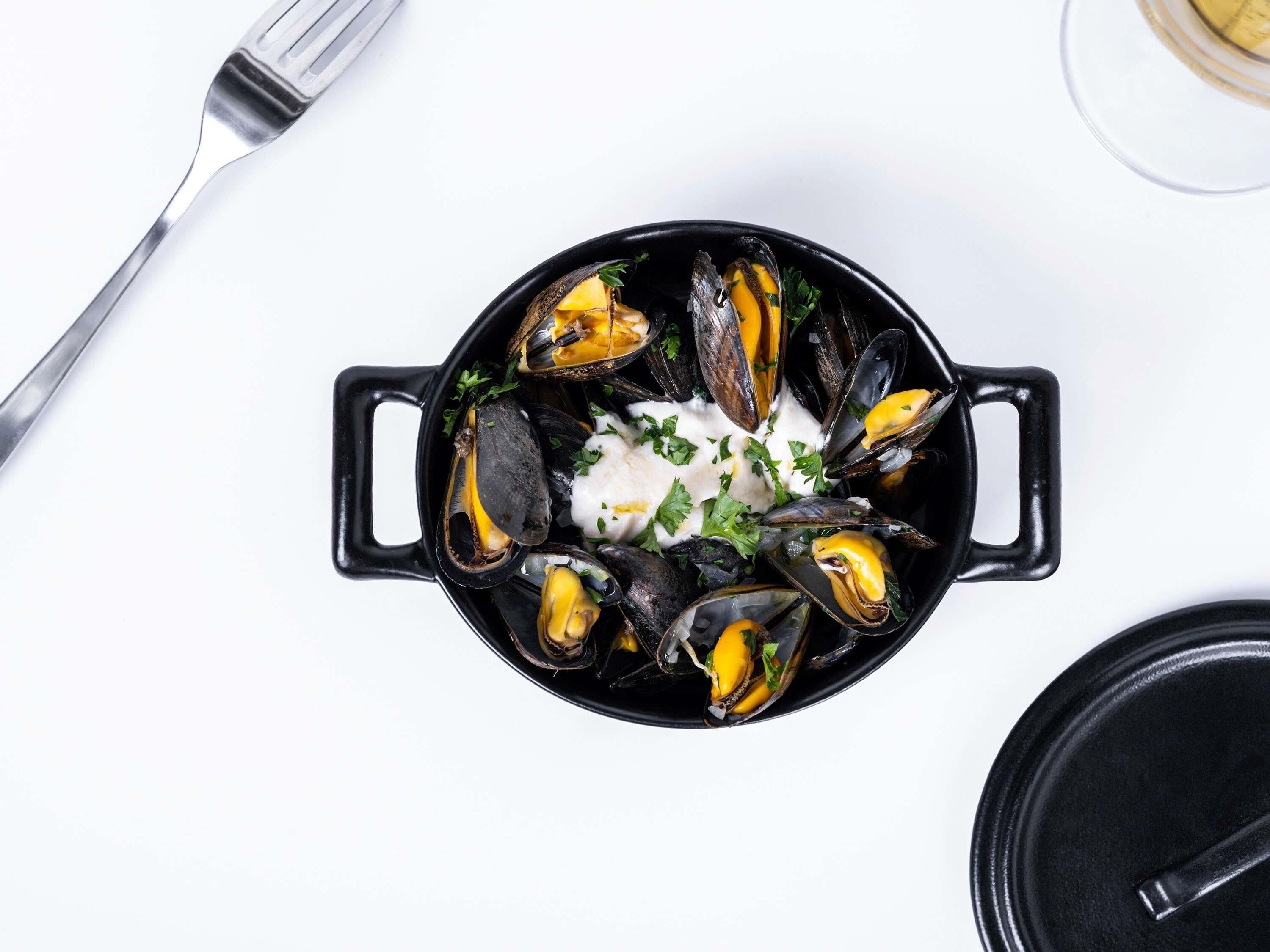 Una olla negra con mejillones y una cobertura cremosa blanca, sobre una superficie blanca.