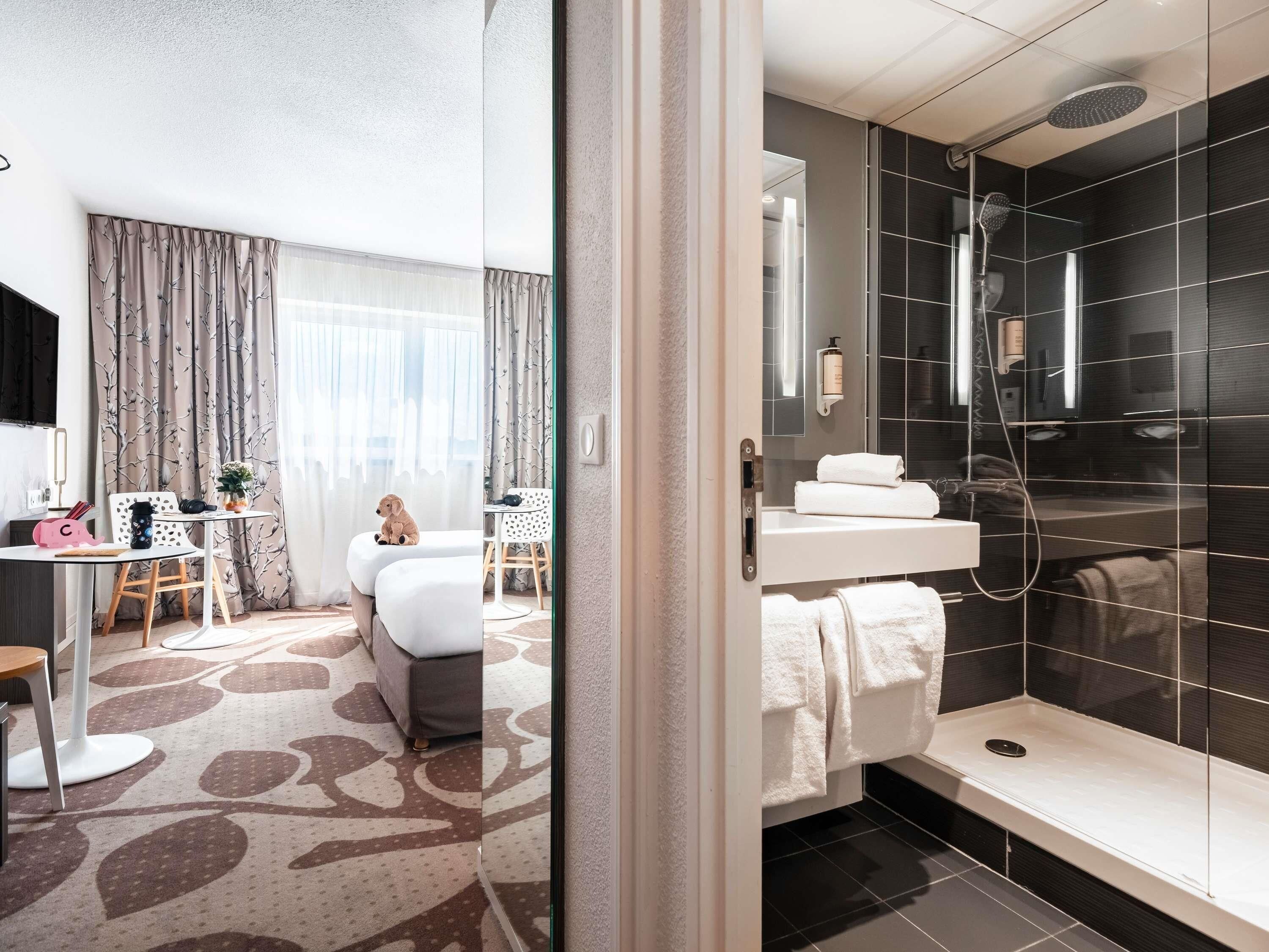 Ett modernt hotellrum med en säng, ett skrivbord, en stol, ett litet bord, en krukväxt, en dusch och ett badrum med spegel och handduksställ.