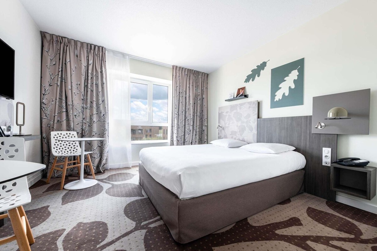 A hotel room with a bed, a desk, a chair, a TV, a window with curtains, and a wall-mounted lamp.