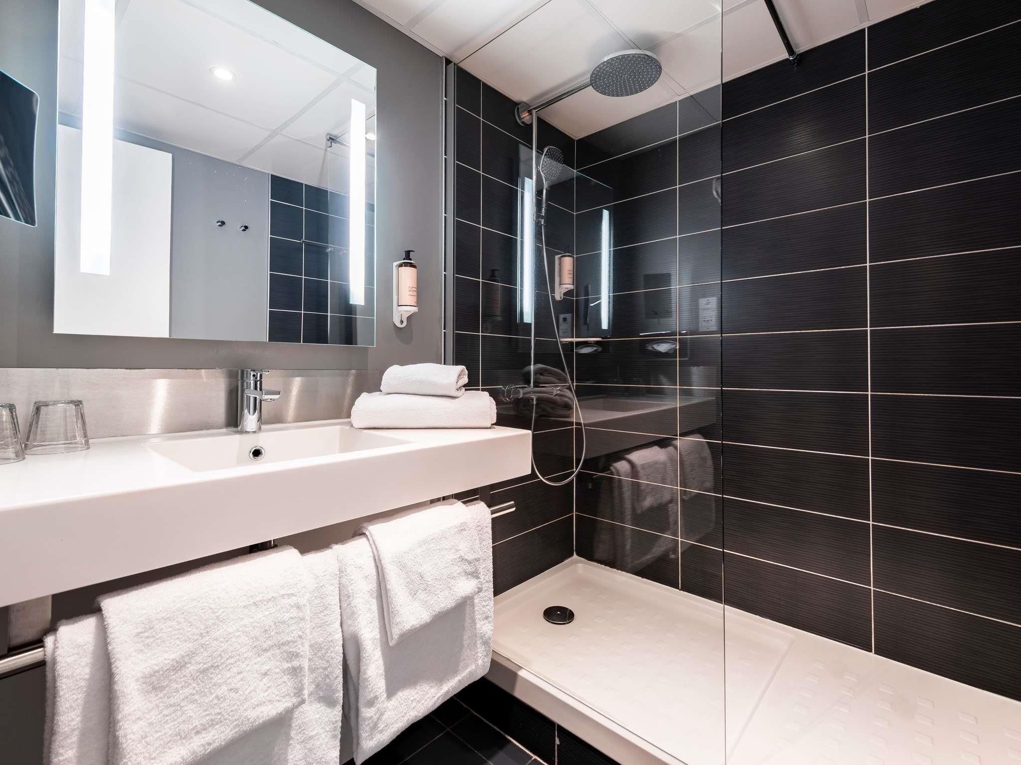 A modern bathroom with a large mirror, a white sink, a shower, and dark tiled walls.