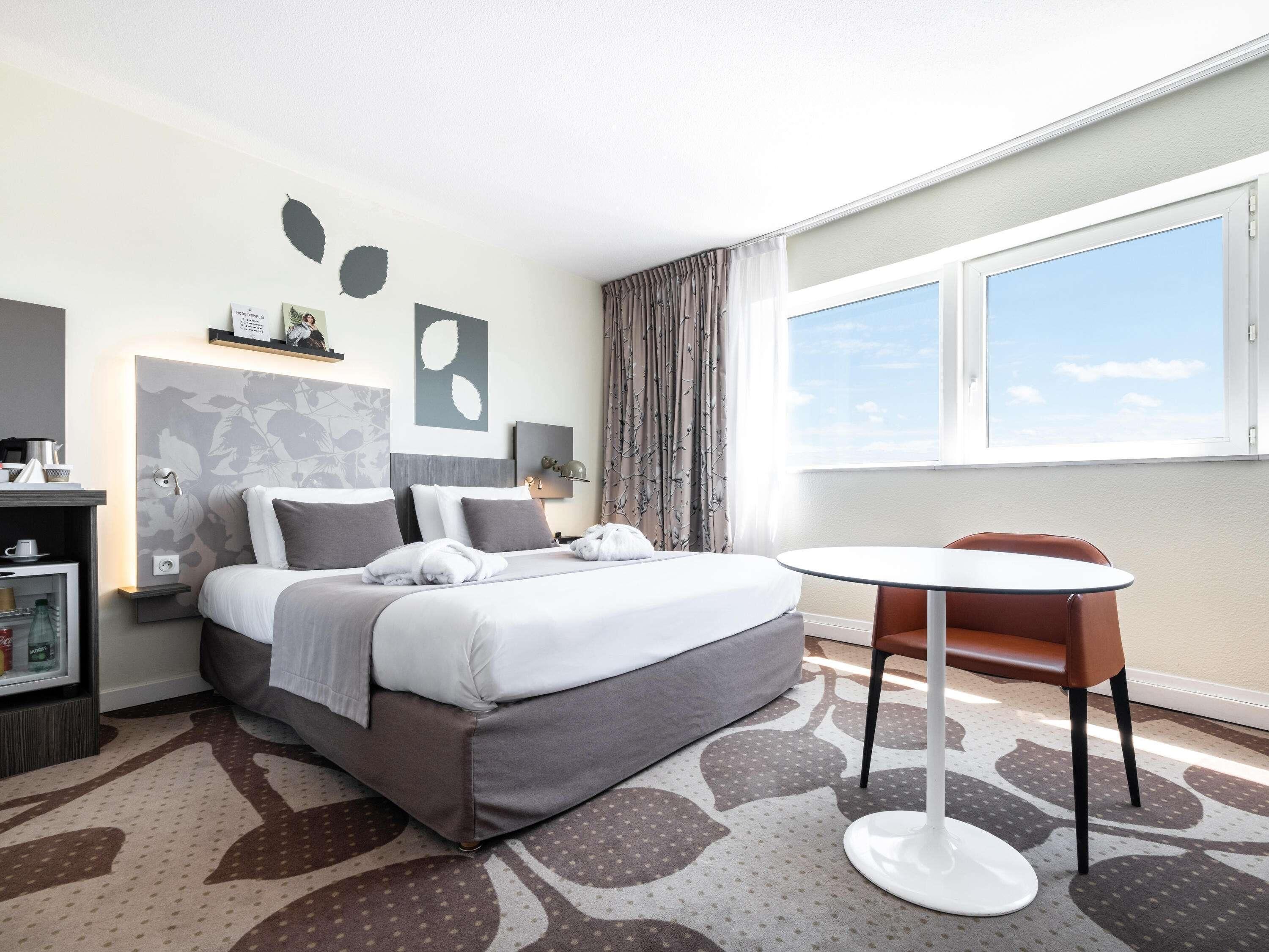 Una habitación de hotel con cama, mesita de noche, silla, ventana y vista al cielo.