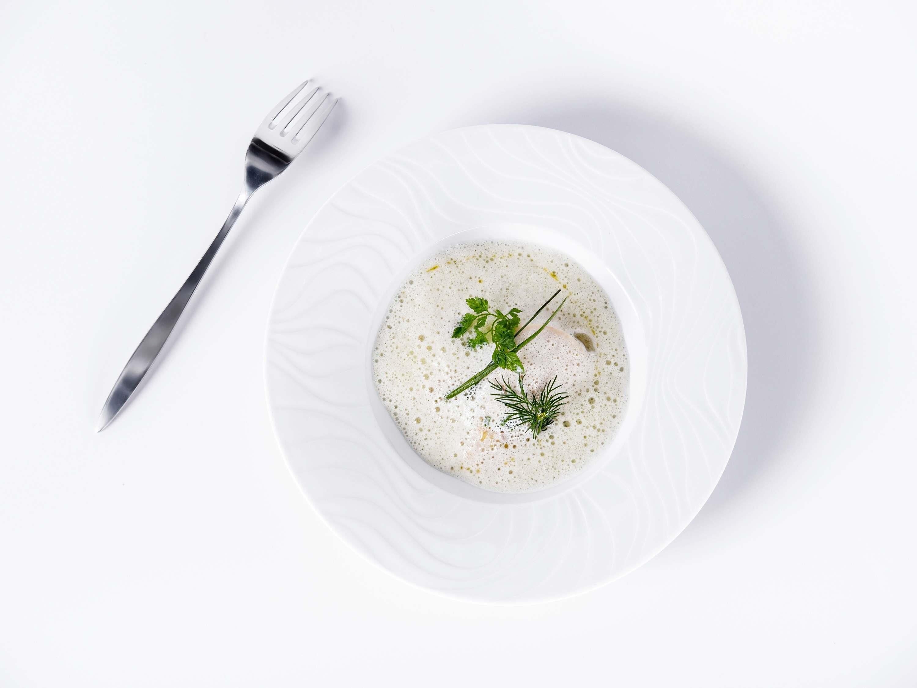 A white plate with a soup garnished with herbs, a fork, and a white textured bowl.