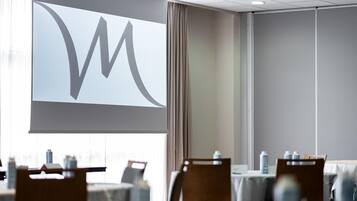 A conference room with a large projection screen displaying a logo, tables with chairs, and a view of buildings outside.