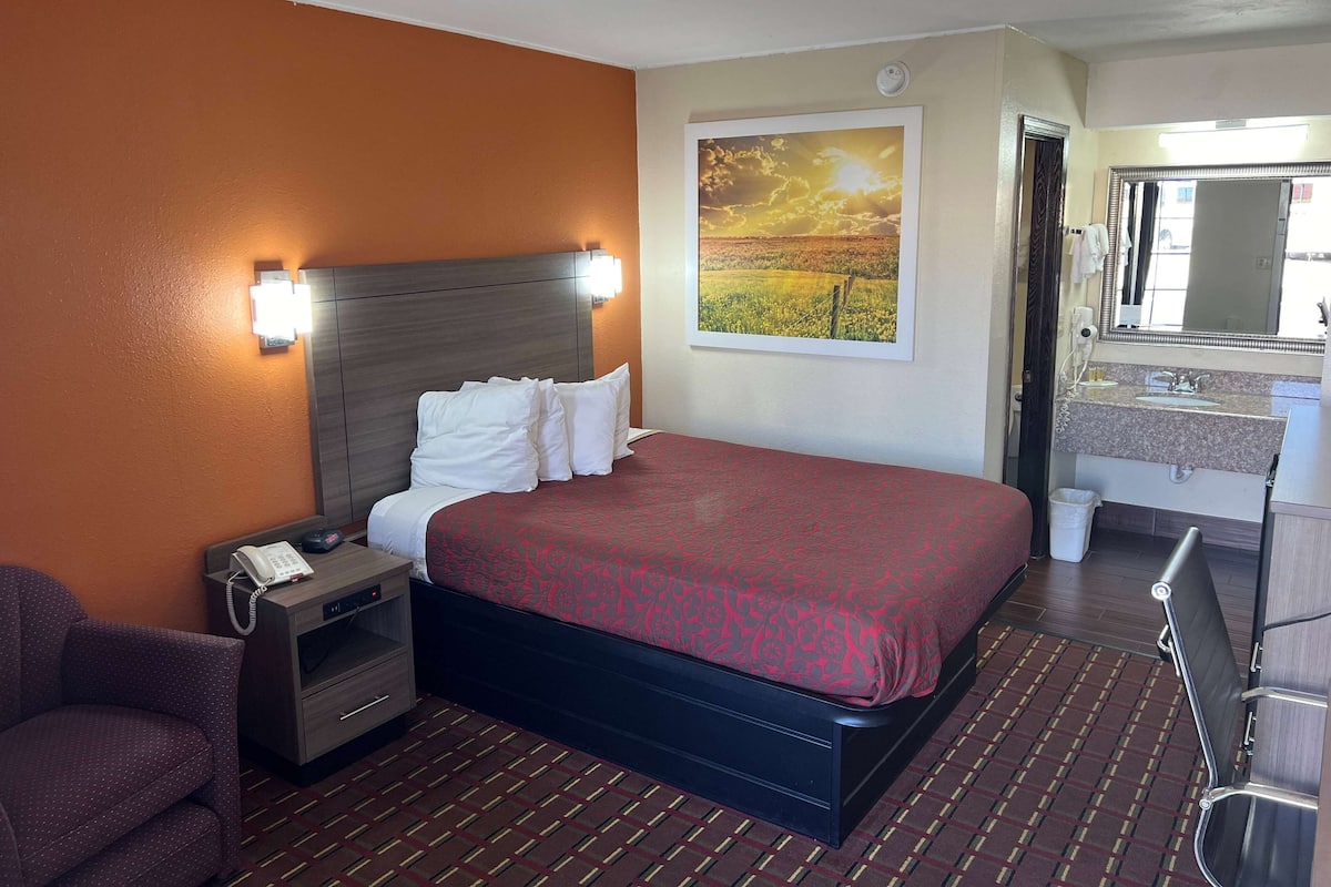 A hotel room with a large bed, a nightstand with a phone, a chair, a bathroom with a sink and mirror, and a painting on the wall.