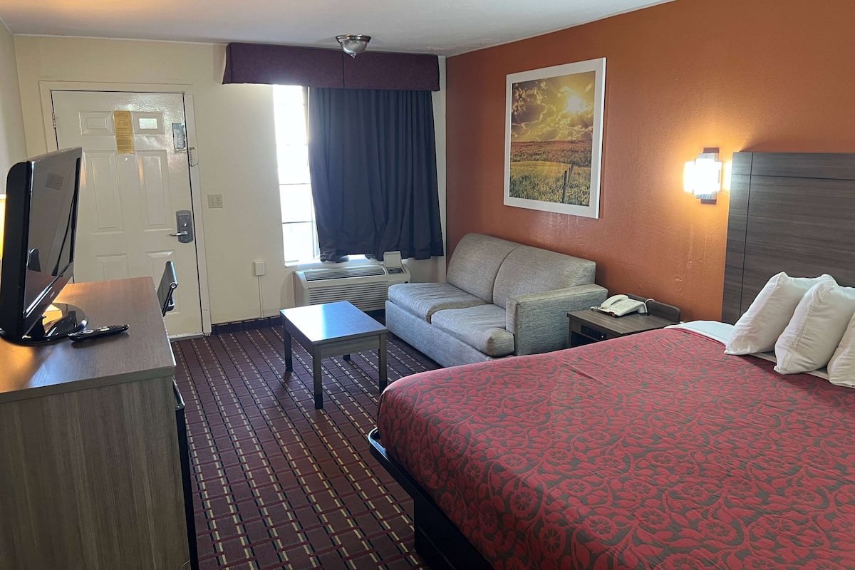 A hotel room with a bed, a desk with a computer, a sofa, a window with curtains, and a wall-mounted light.