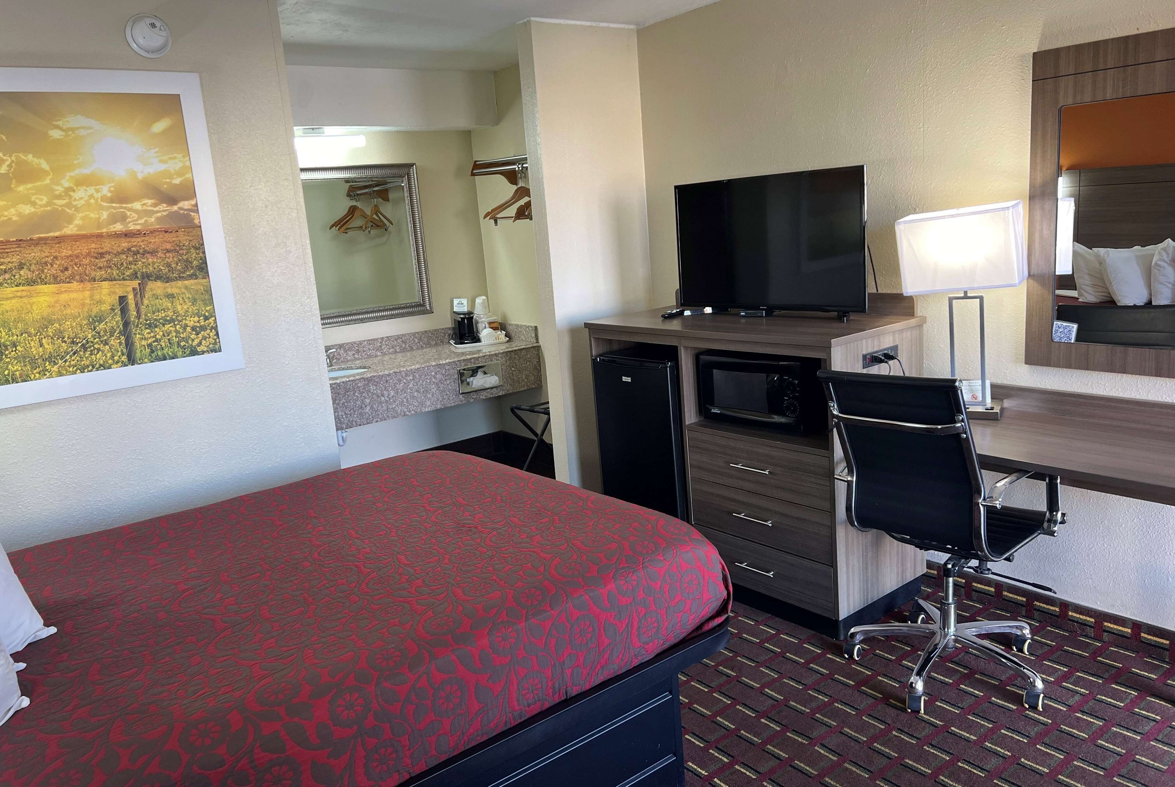 A hotel room with a bed, a desk with a TV, a chair, a desk lamp, a mirror, and a picture of a sunset.