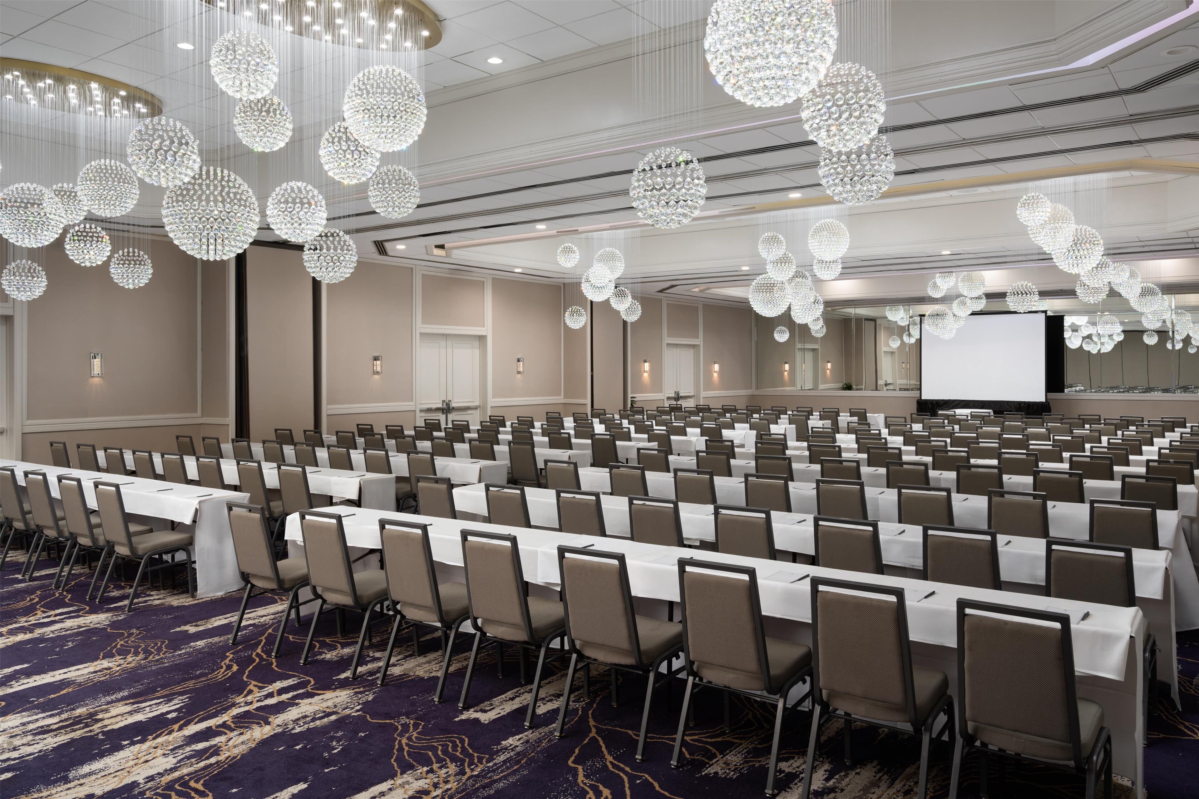 A conference room with rows of tables and chairs, large chandeliers, and a projection screen.