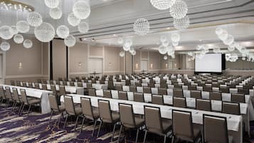 A conference room with rows of tables and chairs, large chandeliers, and a projection screen.