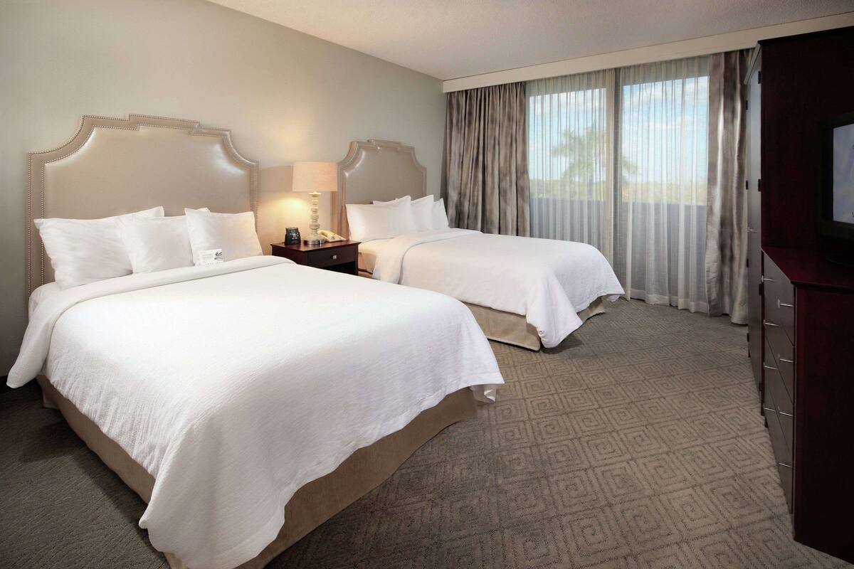 A hotel room with two beds, a TV, a nightstand, and a large window with curtains.