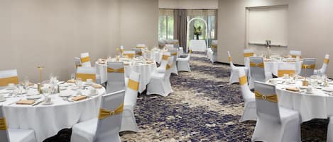 Un salón de conferencias con mesas redondas, sillas blancas y cintas doradas. Las mesas están preparadas con platos, vasos y cubertería. El salón tiene suelo alfombrado y un gran ventanal con cortinas.