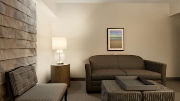 Habitación de hotel con sofá, sillón, mesita de noche con lámpara, cuadro en la pared y un reposapiés estampado.