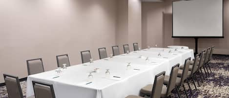 Una sala de conferencias con una larga mesa, sillas, una pantalla de proyector y piso alfombrado.