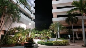 Vista nocturna de un edificio moderno con varios pisos, rodeado de palmeras y un patio empedrado.