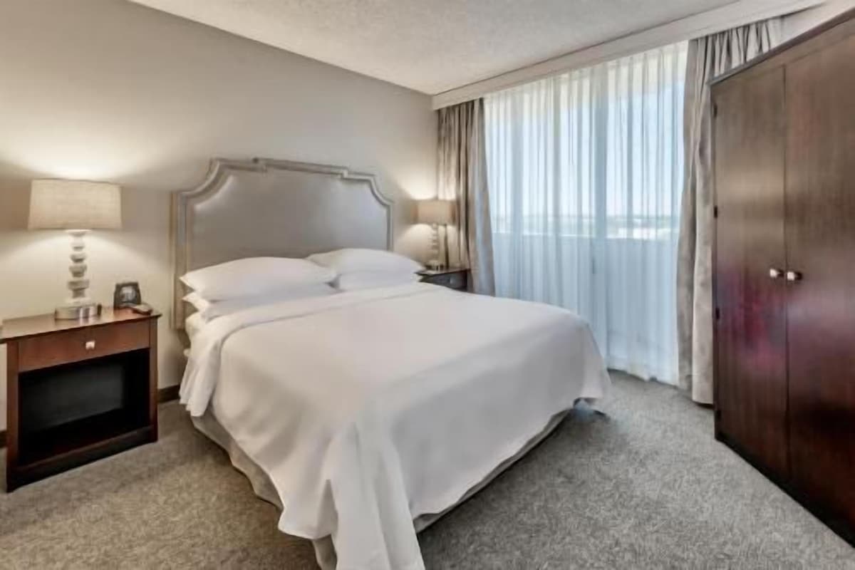 A hotel room with a large bed, a nightstand with a lamp, a wardrobe, and a window with curtains.