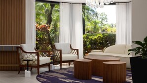 Una sala de estar moderna con dos sillas de madera, un sofá blanco y una mesita de centro. Amplias ventanas con cortinas blancas ofrecen vistas de exuberante vegetación al exterior.