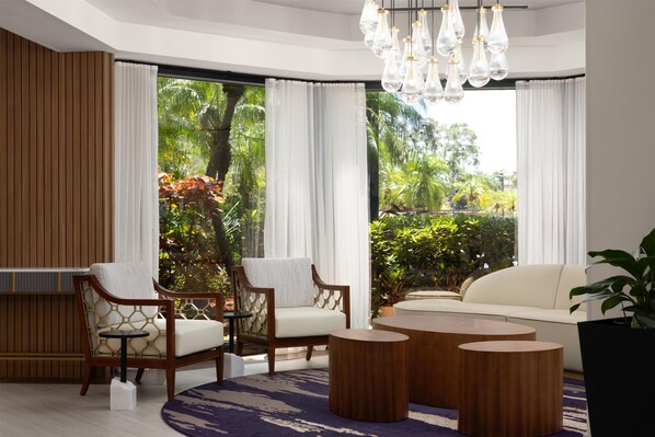 Una sala de estar moderna con dos sillas de madera, un sofá blanco y una mesita de centro. Amplias ventanas con cortinas blancas ofrecen vistas de exuberante vegetación al exterior.