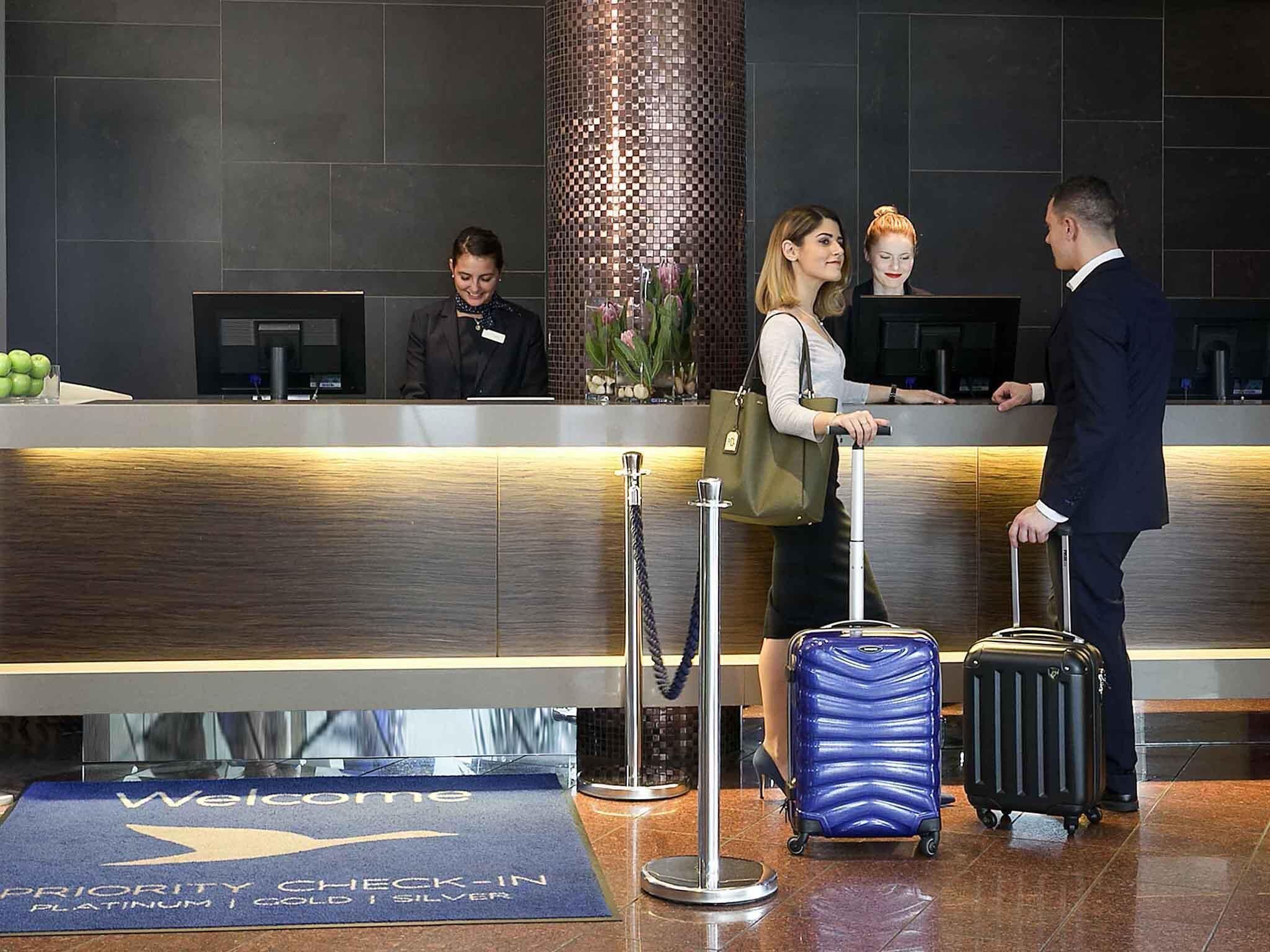 A hotel reception area with a check-in counter, a 'Welcome' mat, and a 'Priority Check-In' sign.