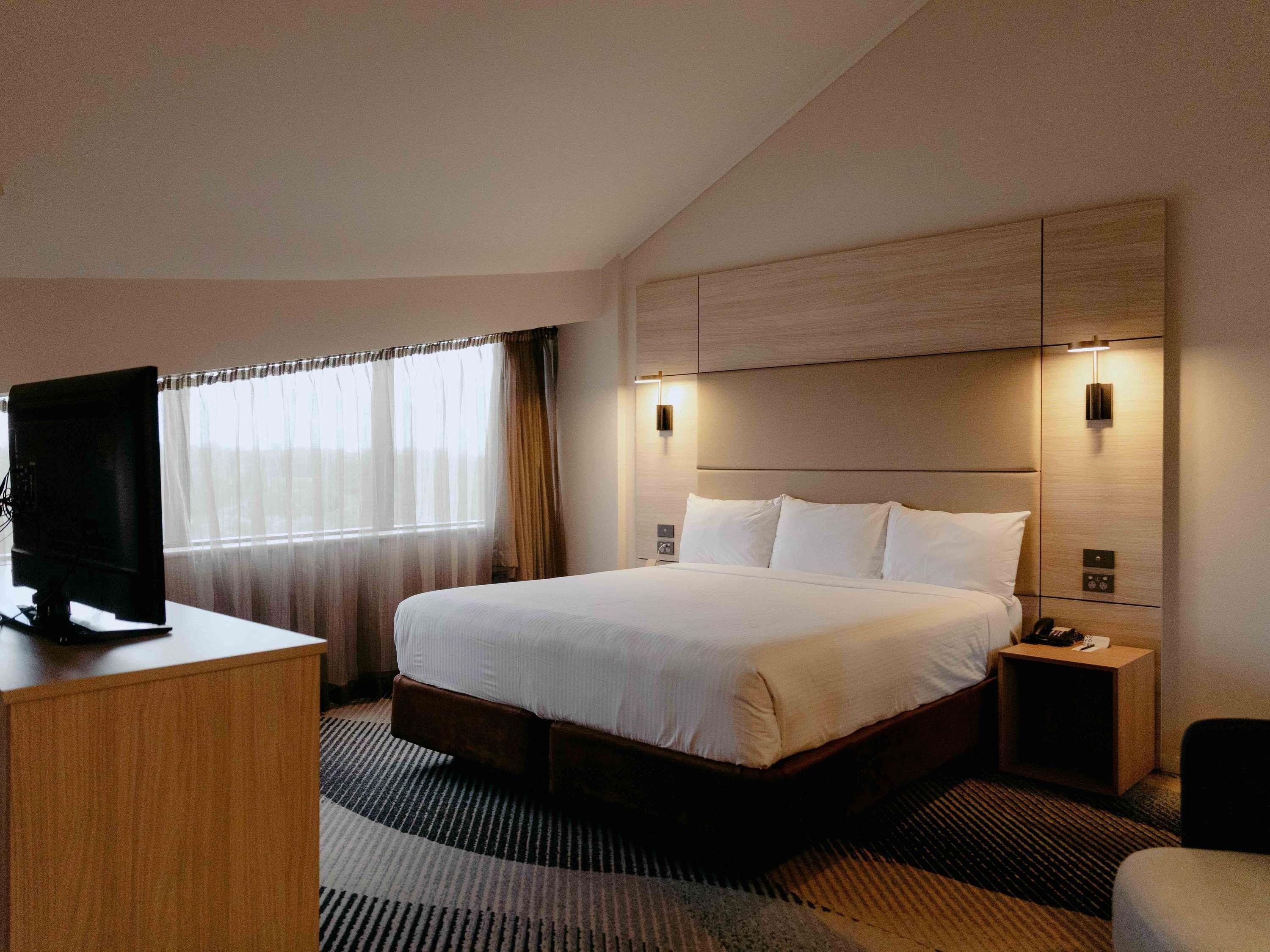 A hotel room with a bed, a desk with a television, a nightstand, and a wall-mounted light fixture.