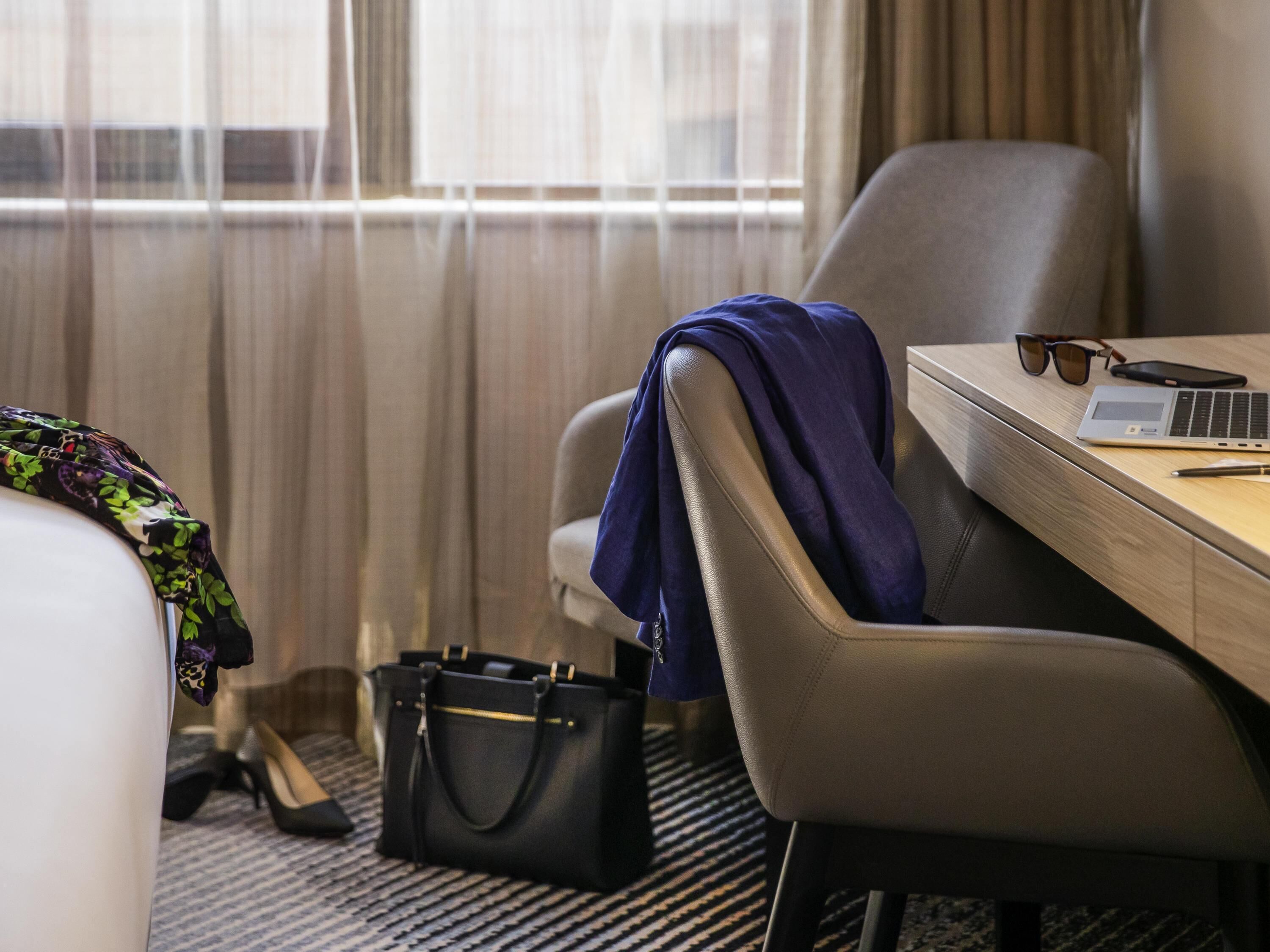 A hotel room with a bed, a desk with a laptop, a chair, a handbag, and a pair of shoes.
