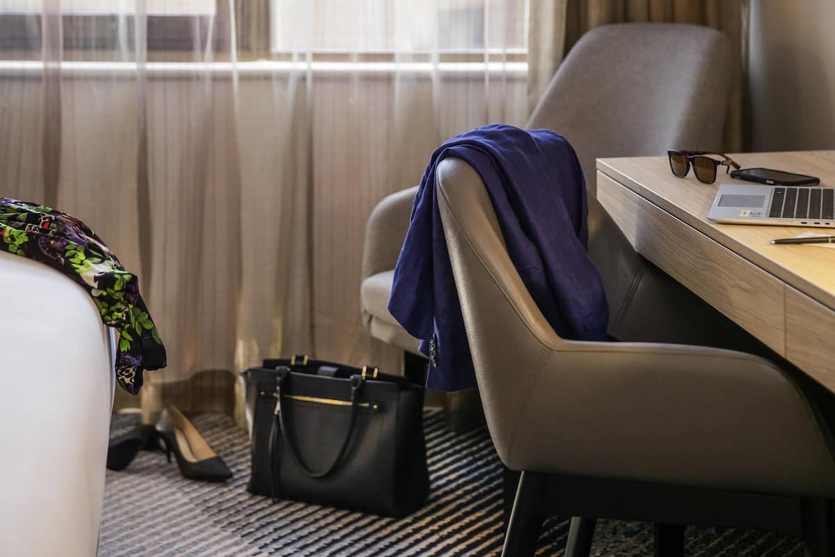 A hotel room with a bed, a desk with a laptop, a chair, a handbag, and a pair of shoes.