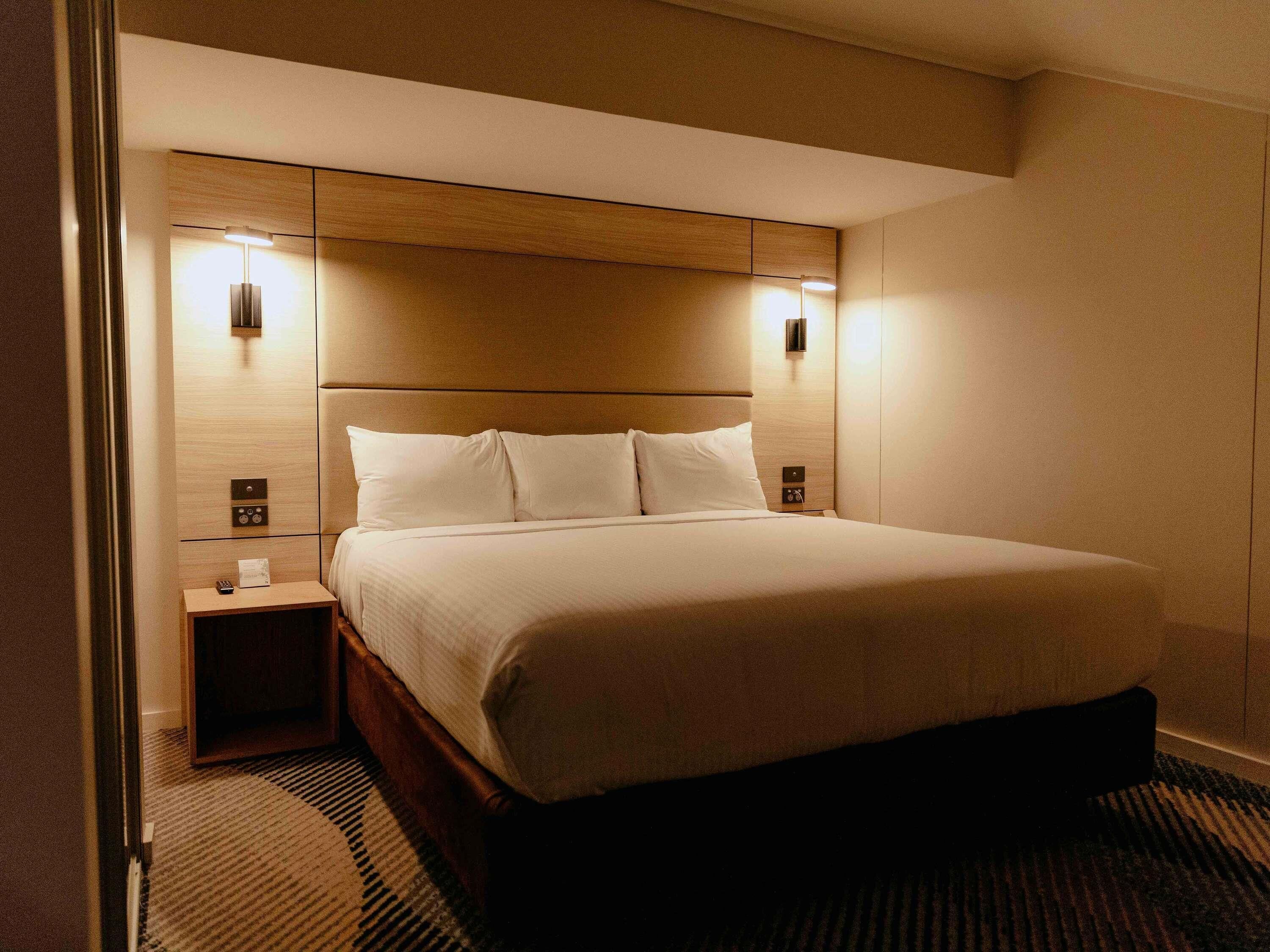 A hotel room with a bed, bedside tables, and wall-mounted lighting.