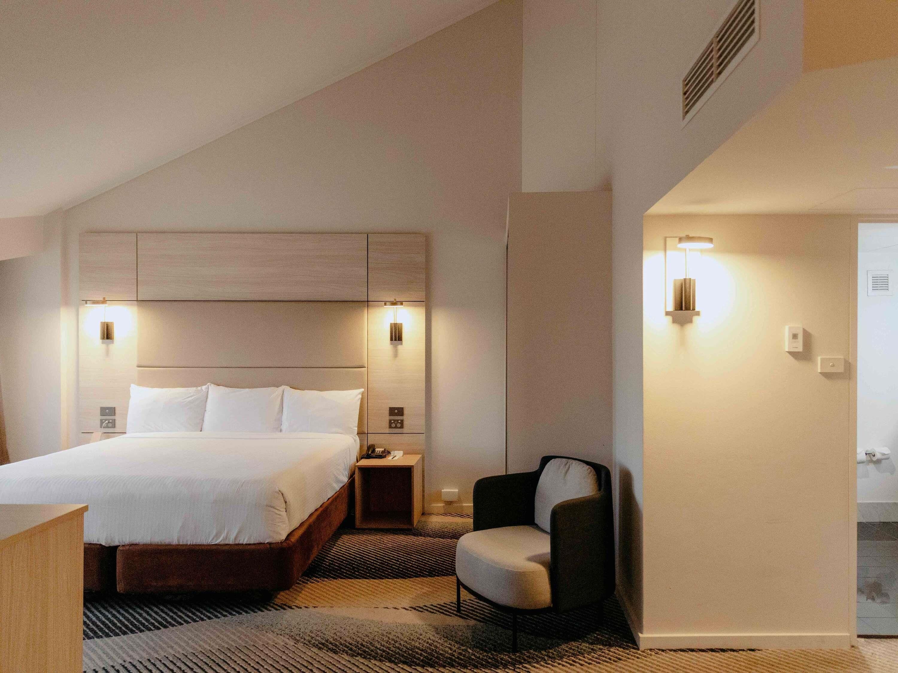 A modern hotel room with a large bed, a chair, a nightstand, and a wall-mounted light fixture.