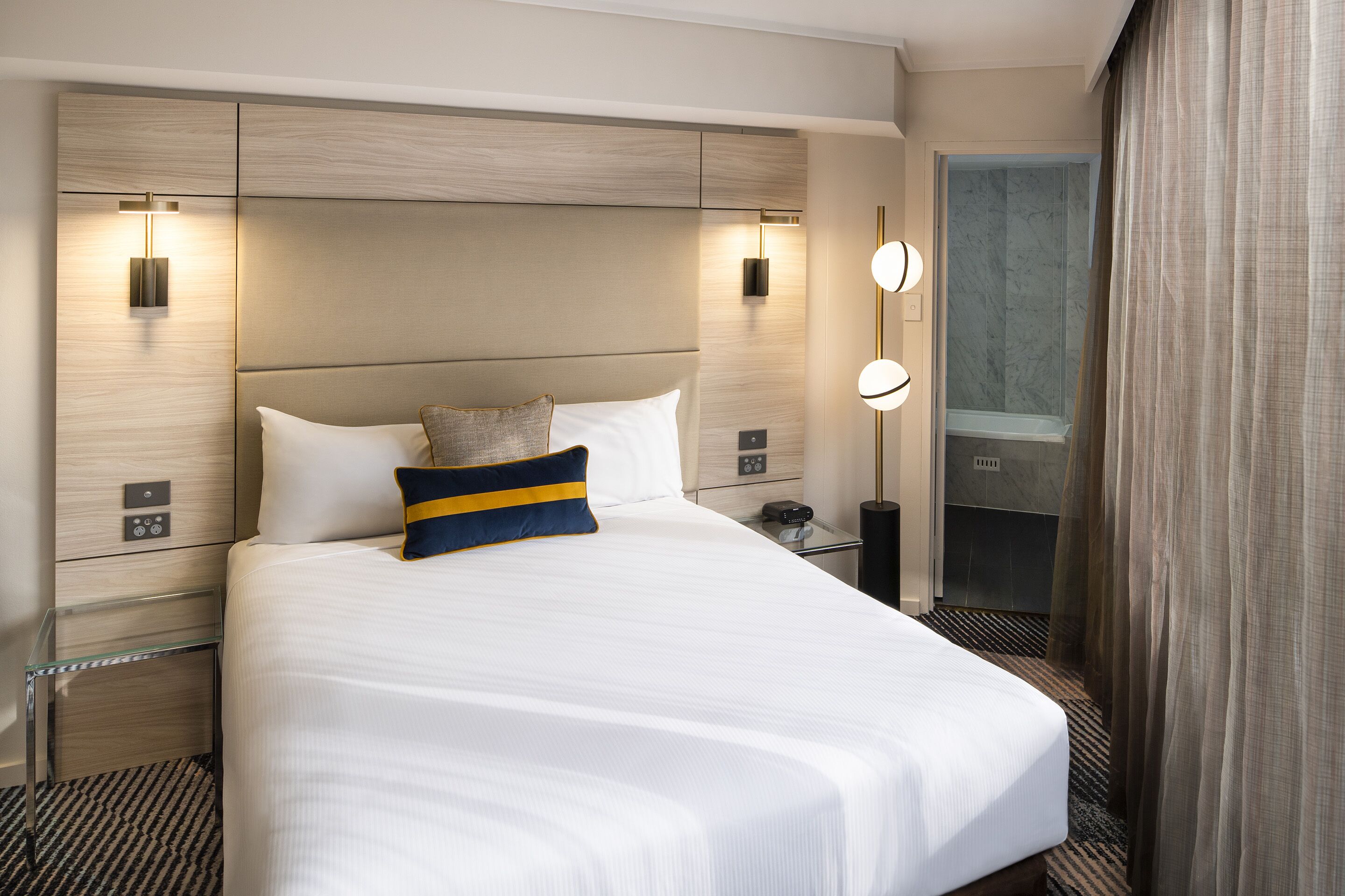 Quarto de hotel com uma cama grande, mesas de cabeceira, espelho e área de chuveiro.