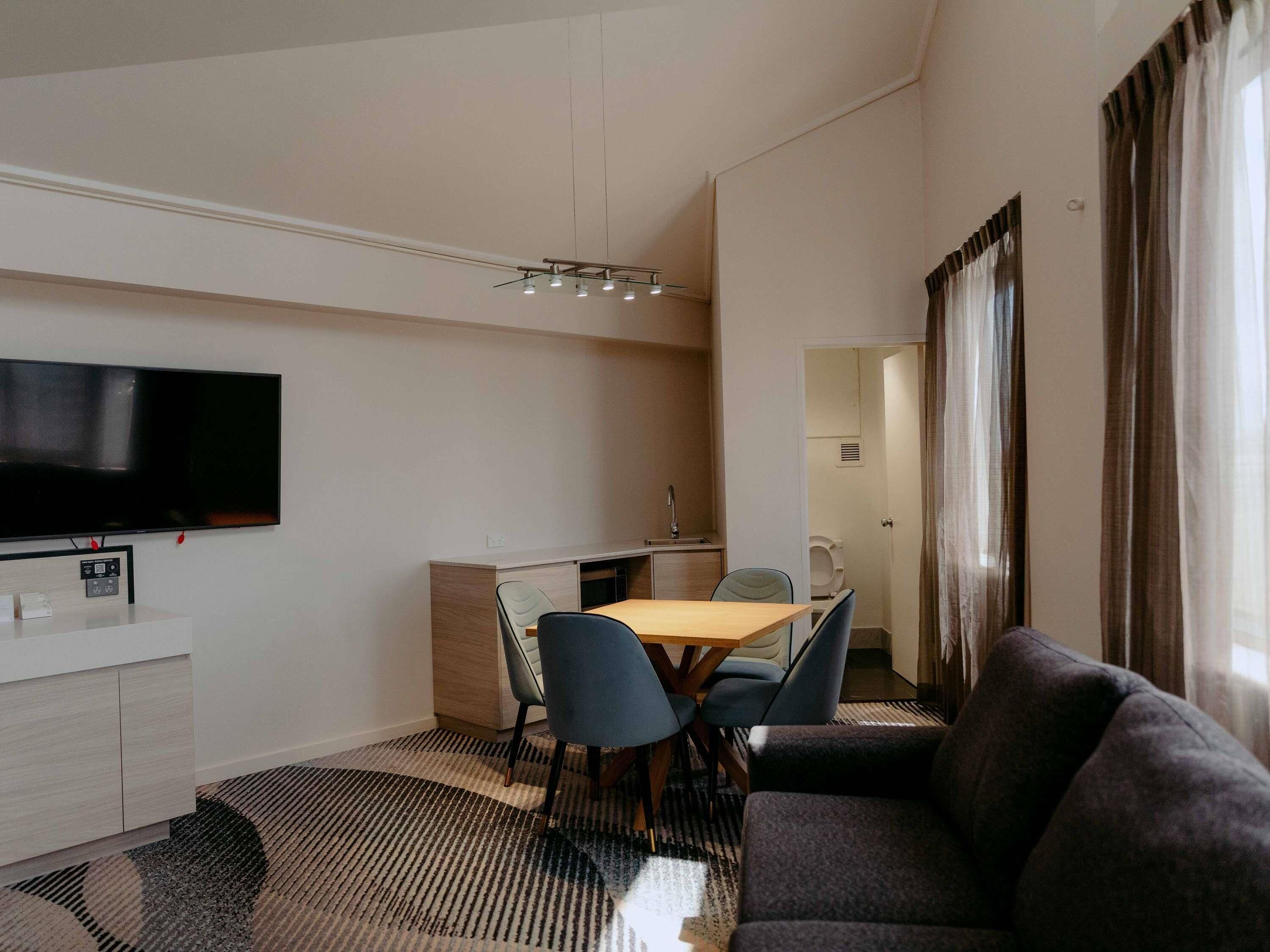 Una habitación de hotel moderna con televisión de pantalla plana, una zona de comedor con mesa y sillas, un sofá y una kitchenette pequeña.
