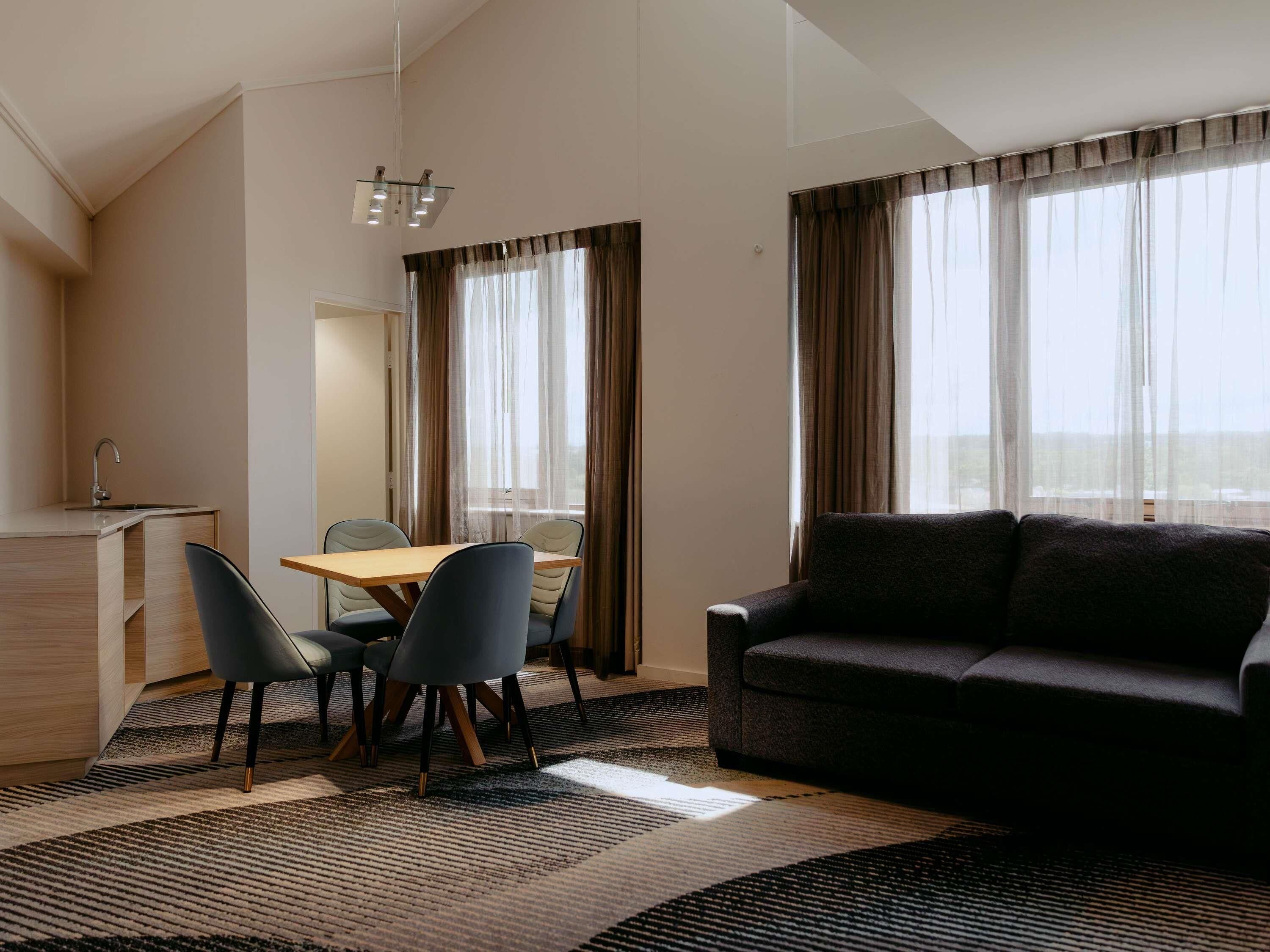A modern hotel room with a dining area, a sofa, and large windows with curtains.