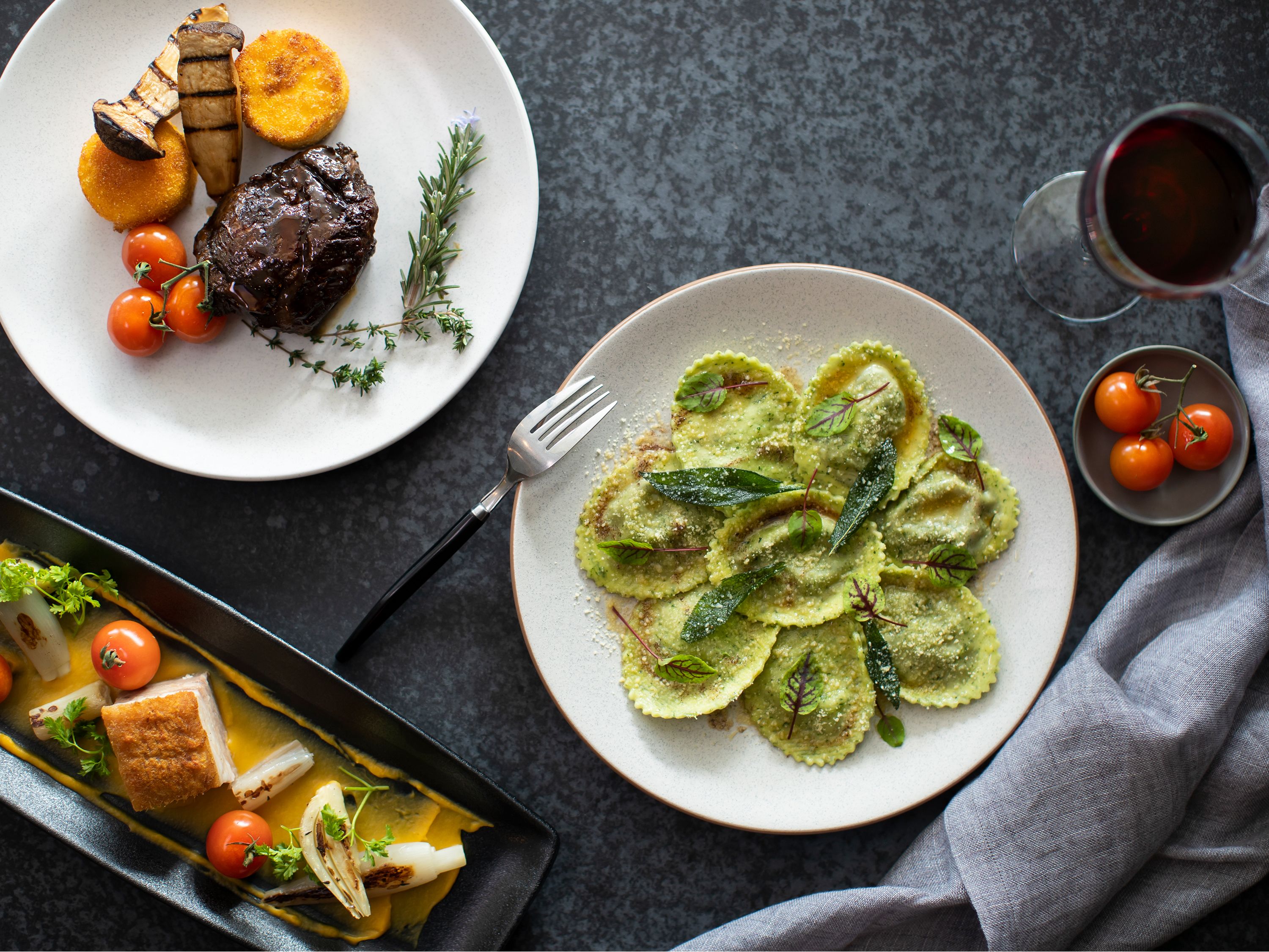 Una mesa de comedor con platos de comida, un tenedor y una copa de vino tinto.