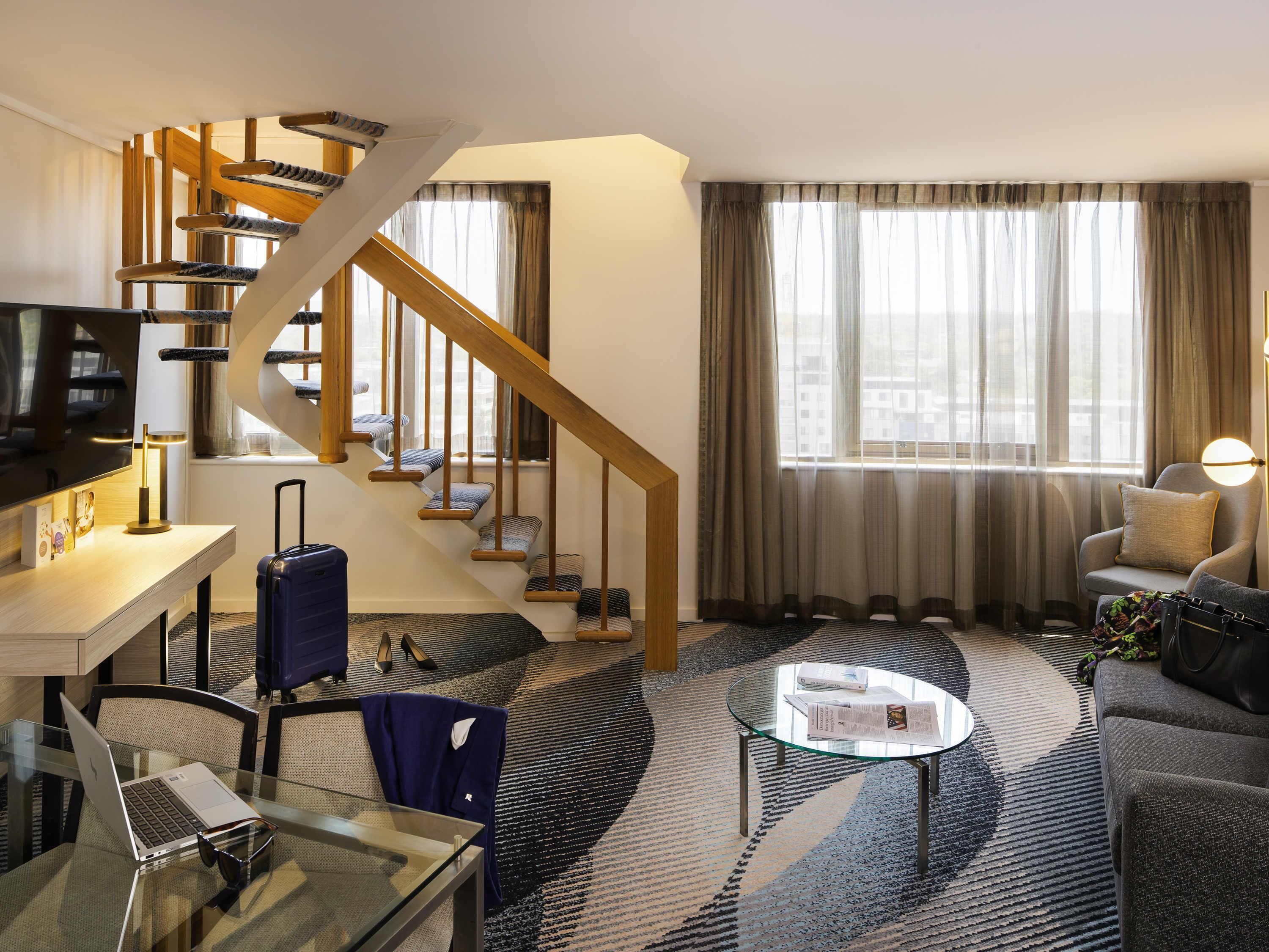 Een hotelkamer met een wenteltrap, een bank, een glazen salontafel, een bureau met een laptop en een televisie.