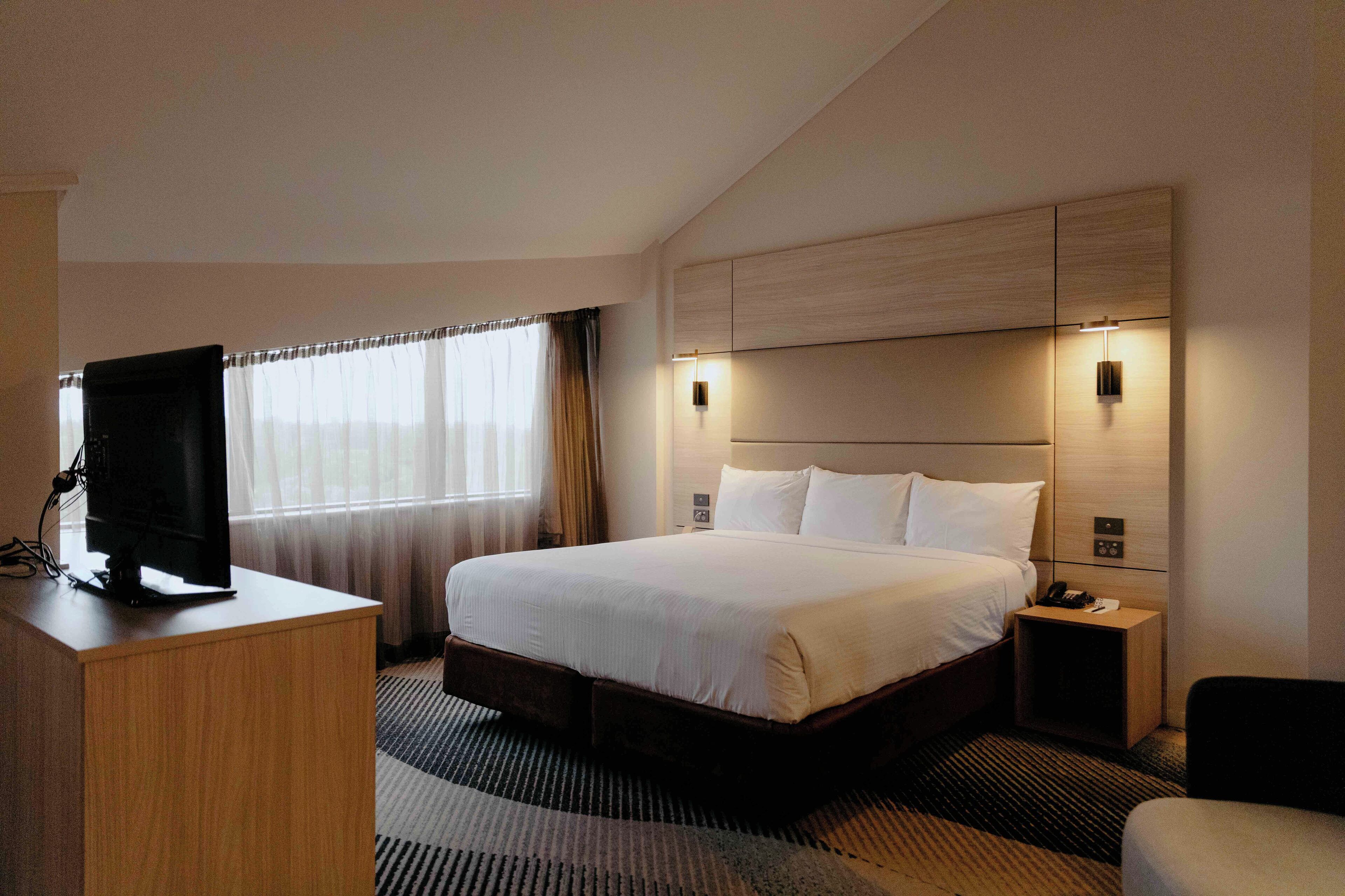 A hotel room with a large bed, a desk with a television, a bedside table, and a wall-mounted light fixture.