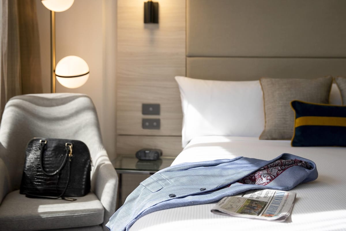 Una habitación de hotel con una cama, una silla, un bolso y una lámpara.
