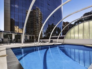 A swimming pool with a glass enclosure, outdoor seating, and a modern building in the background. - Novotel Sydney Parramatta (Parramatta)