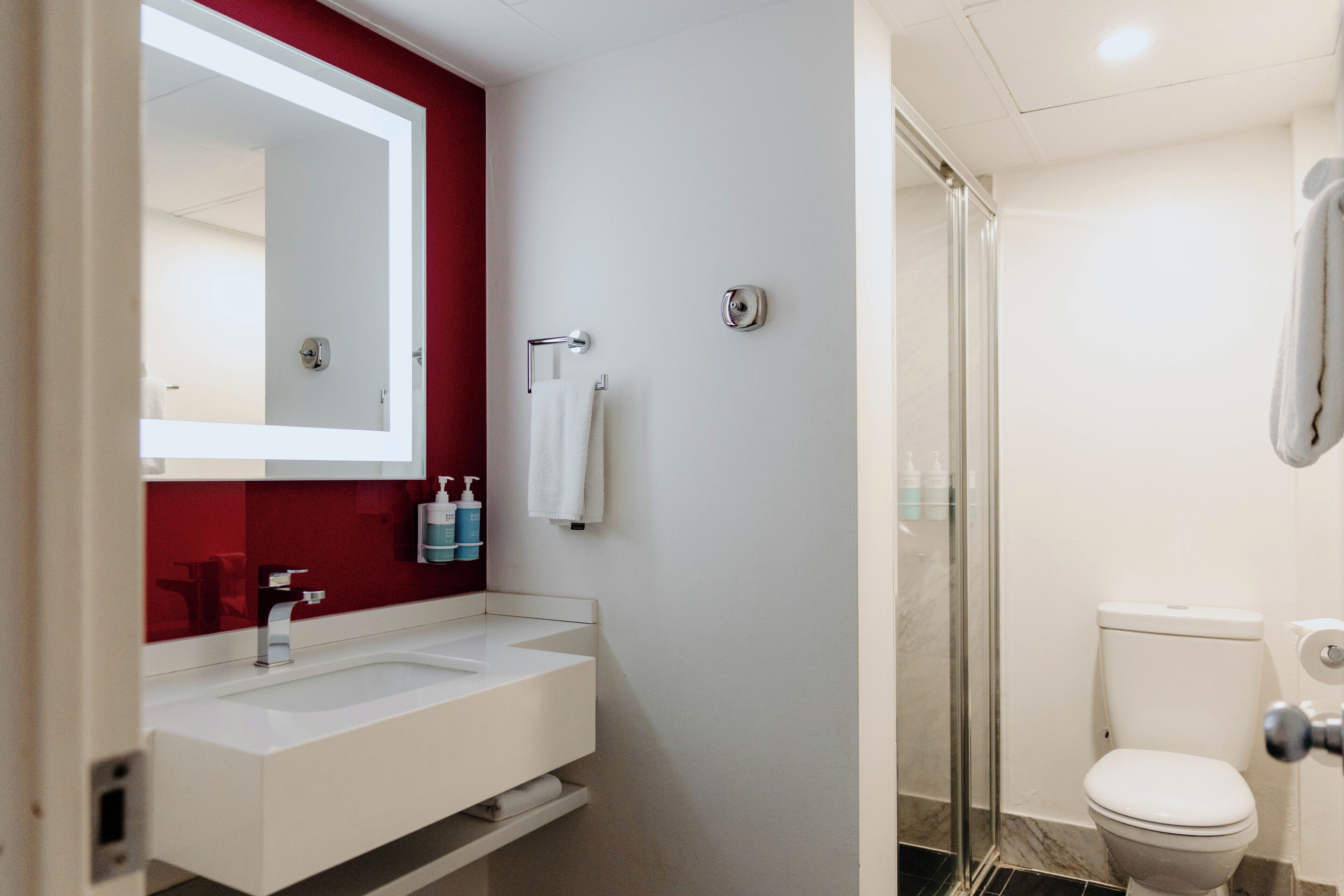 Une salle de bain moderne avec un lavabo blanc, un mur d’accent rouge, une douche en verre et des toilettes.